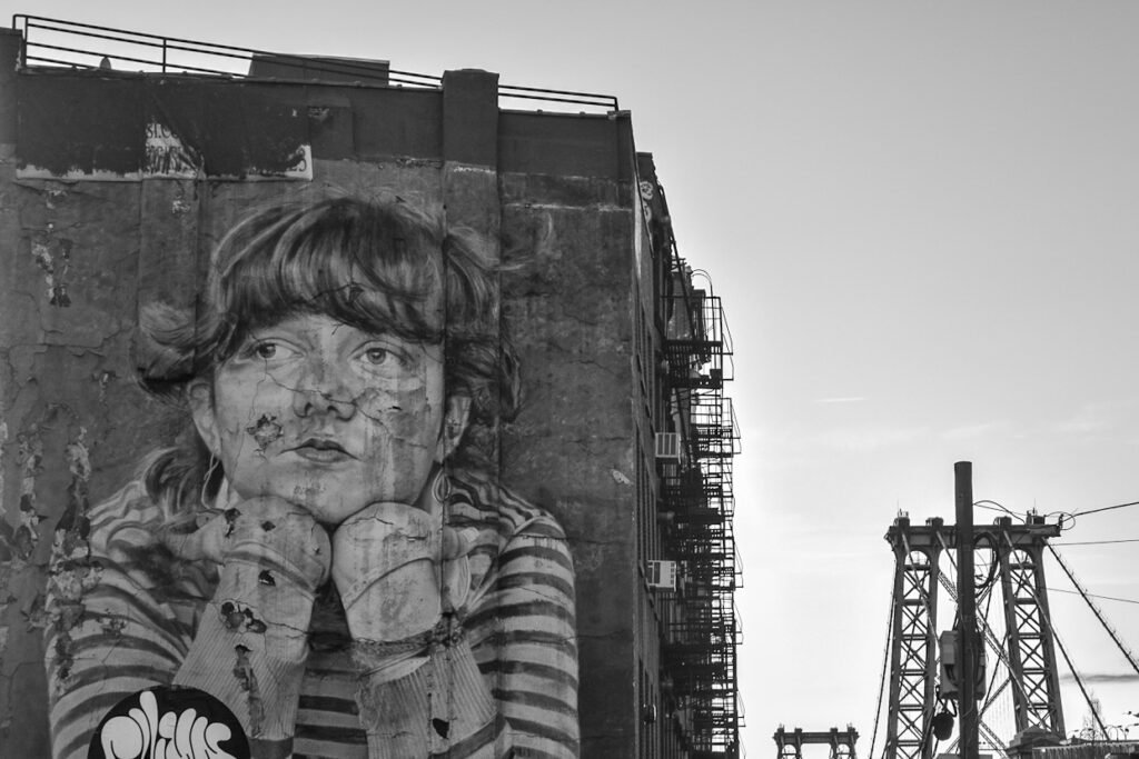 A dramatic black and white image of a large-scale mural or graffiti of a girl painted on a cracked brick wall. The wall is adjacent to an industrial building with a fire escape. A white quote is overlaid on the image, reading: "The creative act is not about inventing something new, but about revealing what has always."
