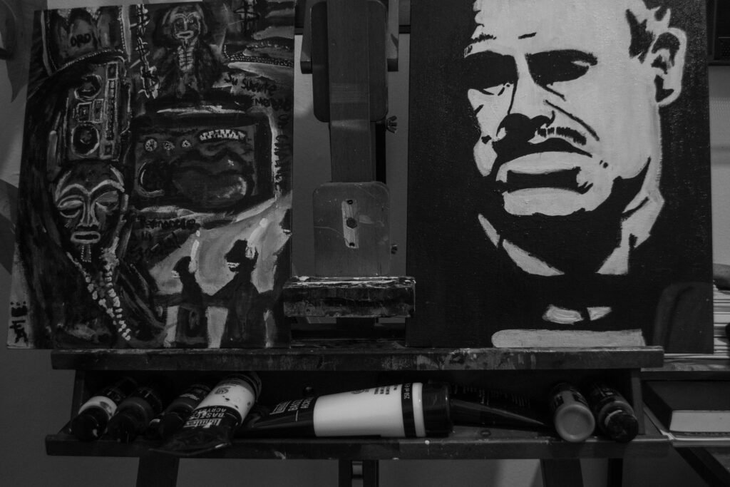 "THE DANCE OF THE HANDS": A black and white photo of an artist's easel in a studio. The main canvas is a high-contrast stencil portrait of a man. Tubes of paint and other art supplies are visible at the base of the easel.