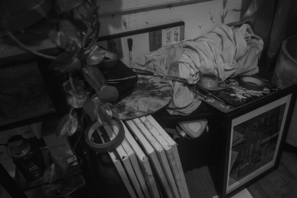 "THE SOUL'S RESONANCE": A dark, black and white shot of a cluttered corner, possibly an art studio, showing various objects, canvases, and what appear to be stacked, organic-looking structures or sculptures.
