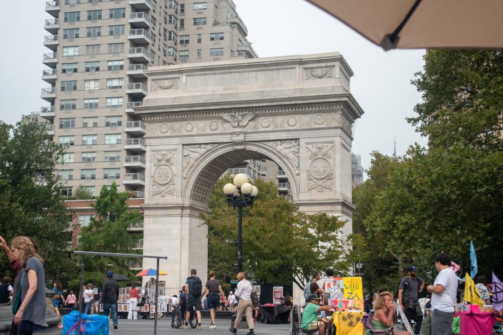 Washington Square Arch: A mid-shot view of the Washington Square Arch in New York City, with artists and art vendors gathered near the base of the arch and people walking around.