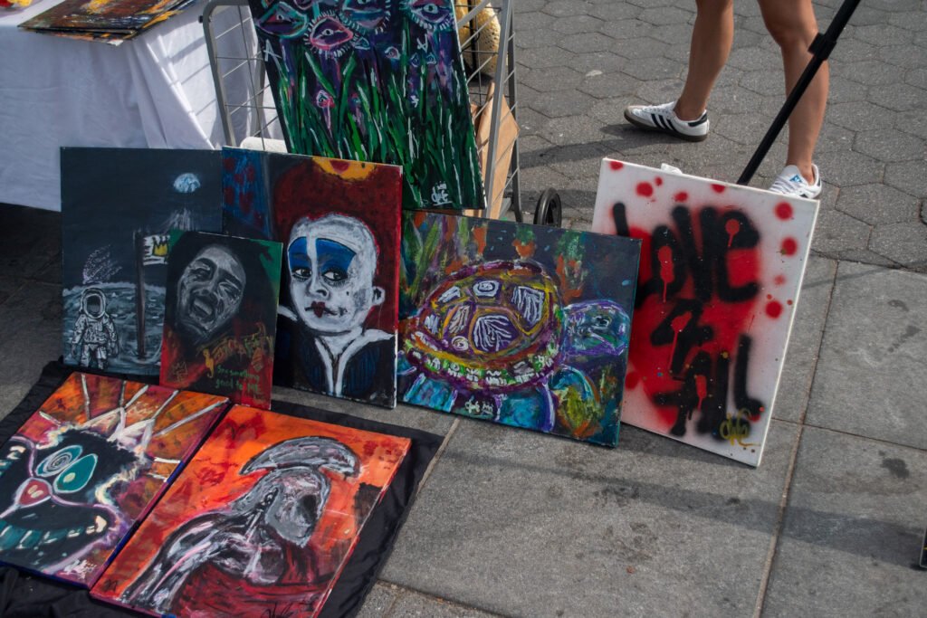 Three canvases in an urban, pop-art style displayed directly on the pavement. The paintings feature vibrant colors, abstract figures, and skull imagery.