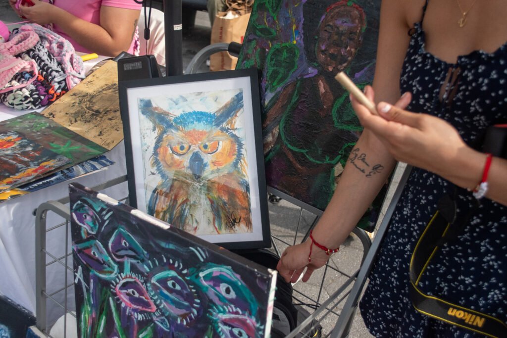A close-up of a vendor in a floral dress holding an incense stick or piece of chalk near an easel. A folk-art style painting of an owl and a canvas with a monster image are clearly visible in the foreground.