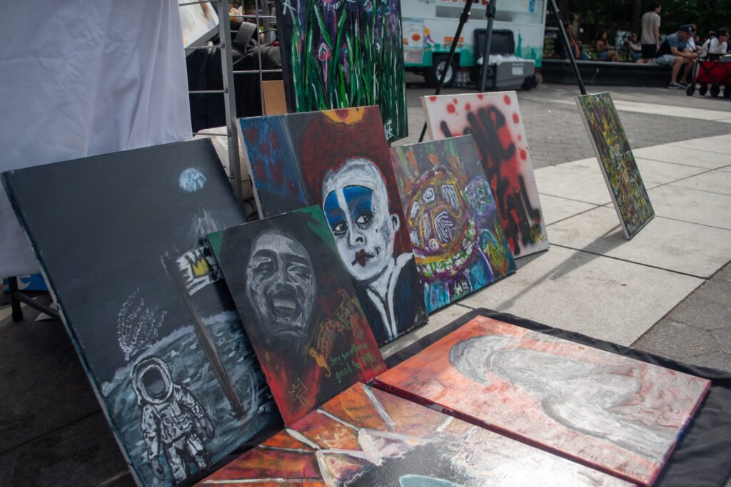 A row of large canvases featuring dark, expressionistic street art, including skull portraits, ghostly faces, and abstract figures, laid out on the ground.
