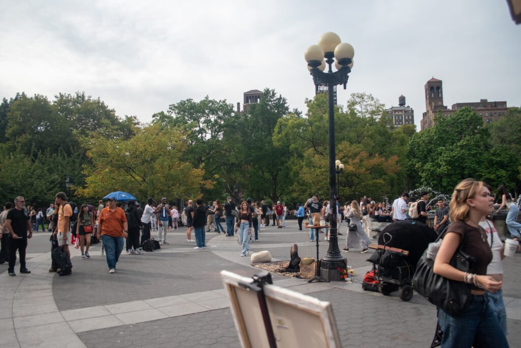 A view of a wide pathway in Washington Square Park filled with people, with lush trees on the left and a tall lamppost on the right. An artist or vendor is visible in the right foreground.